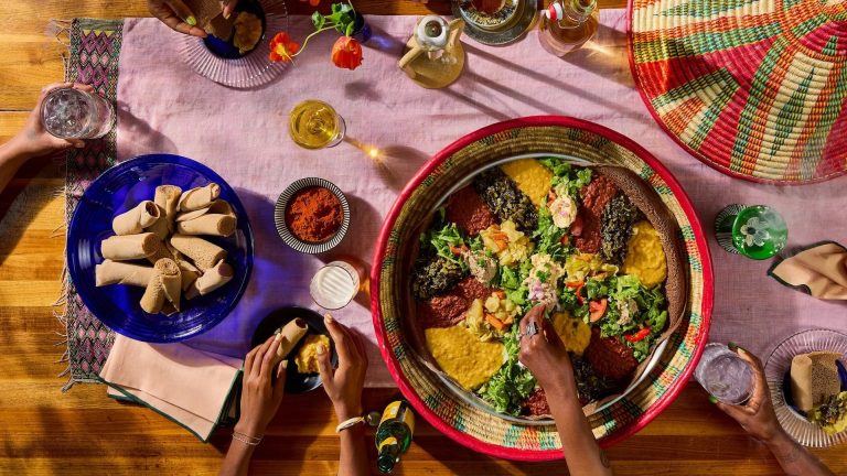 Overhead view of a combination platter of Ethiopian stews, greens and salads on injera in a woven tray, with rolled injera, a bowl of berbere and drinks on a table