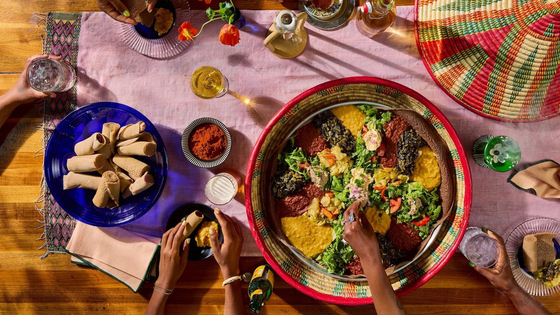 Overhead view of a combination platter of Ethiopian stews, greens and salads on injera in a woven tray, with rolled injera, a bowl of berbere and drinks on a table